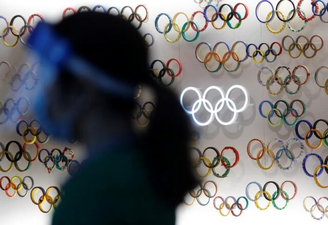 pessoa-com-mascara-e-face-shield-em-frente-a-simbolos-das-olimpiadas-em-toquio